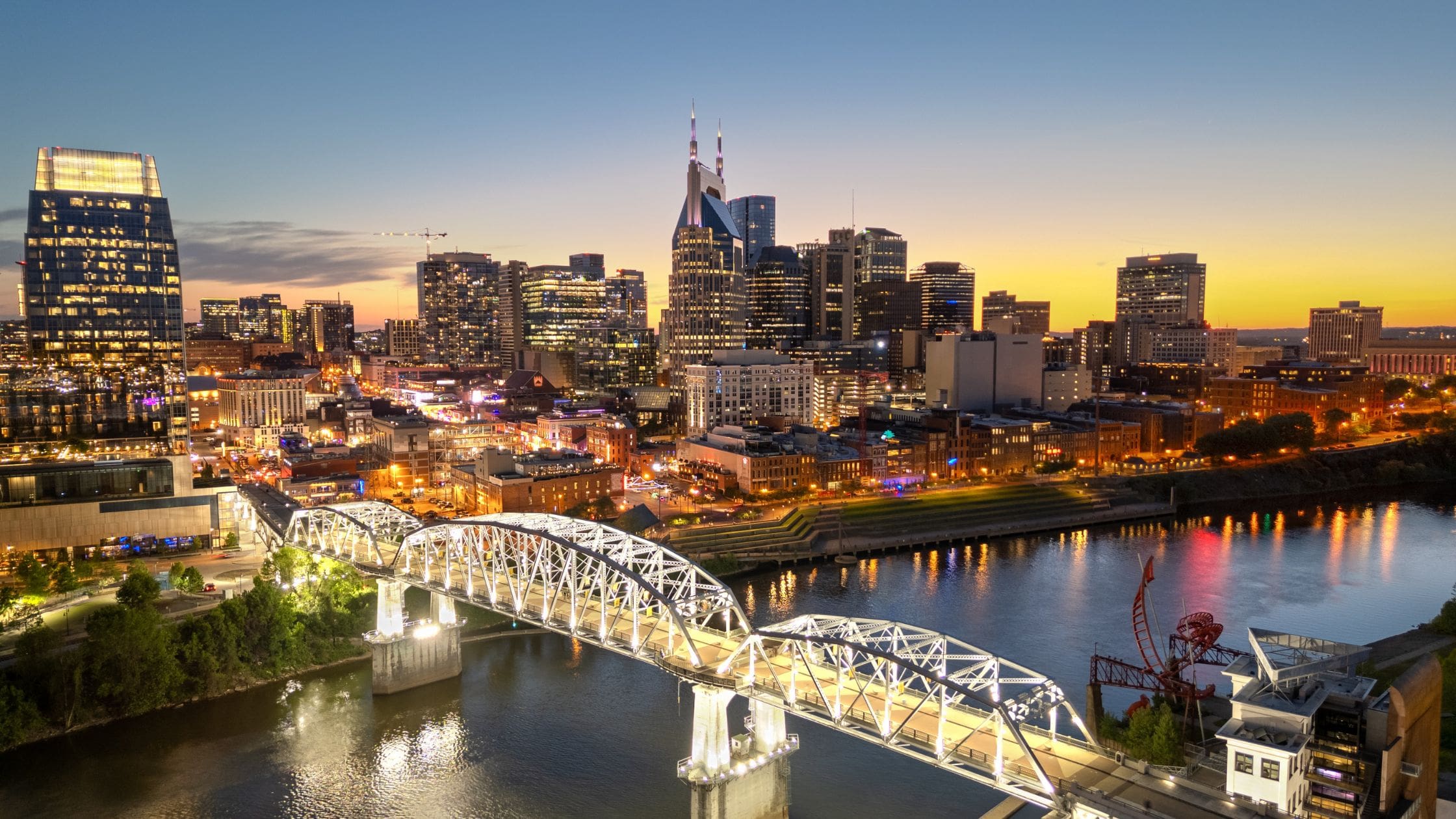Downtown Nashville skyline at dusk seen across the Cumberland River, with the John Seigenthaler Pedestrian Bridge in the foreground